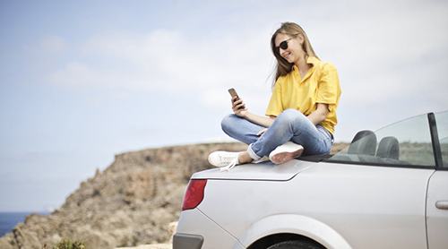 Lady sitting at the back of her car holding a cellphone - Free Insurance Quotes